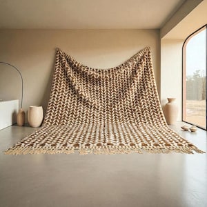 May include: A large, brown and white patterned blanket with a fringed edge is draped against a wall. The blanket features a repeating chevron design. The room has neutral tones, with a large window and decorative vases.