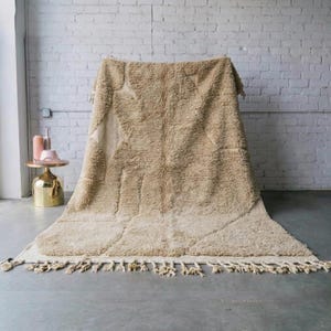 Op de afbeelding: Een groot, beige Marokkaans tapijt met een geometrisch patroon en franjes. Het tapijt is gemaakt van een dik, getextureerd materiaal en wordt gepresenteerd tegen een witte bakstenen muur. De afmetingen van het tapijt zijn niet zichtbaar.