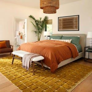 Large handmade Moroccan high-pile shag rug in saturated Thai curry mustard-gold, displayed vertically against a white brick wall. Surface features a tone-on-tone square grid pattern formed through pile variation — raised mustard squares separated by recessed ivory cream channel lines across the entire field. Dense, plush, curly wool pile texture throughout. Knotted braided ivory fringe along the top hem edge and long hand-tied ivory fringe along the bottom edge.