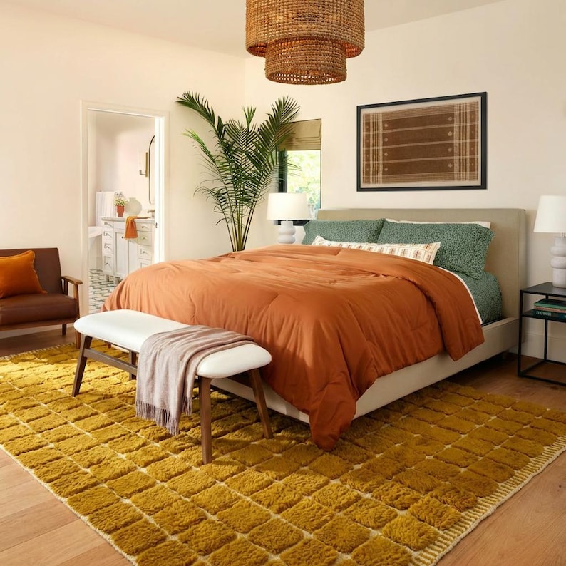 Large handmade Moroccan high-pile shag rug in saturated Thai curry mustard-gold, displayed vertically against a white brick wall. Surface features a tone-on-tone square grid pattern formed through pile variation — raised mustard squares separated by recessed ivory cream channel lines across the entire field. Dense, plush, curly wool pile texture throughout. Knotted braided ivory fringe along the top hem edge and long hand-tied ivory fringe along the bottom edge.