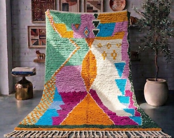 Può includere: Un tappeto rettangolare a pelo lungo caratterizzato da un vivace motivo geometrico nei toni del verde, giallo, magenta, blu e crema. Il design include forme triangolari e astratte, rifinito con frange color crema sul bordo inferiore.