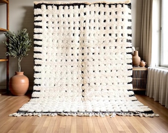 May include: A large, rectangular rug with a cream background and a repeating pattern of black squares and lines. The rug has a thick, textured pile and a fringe along the bottom edge. The rug is displayed on a wooden floor.