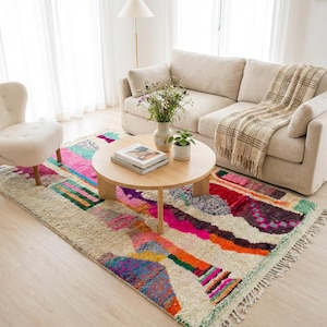 Colorful Moroccan wool rug in a boho living room, featuring an ivory base with abstract patchwork blocks in pink, orange, green, burgundy, black, yellow, and mint, plus playful linear motifs and braided ivory fringes. The rug has a plush high pile and hand-knotted texture, styled under a round rattan coffee table with a cream sofa, earthy pillows, woven pouf, plants, macramé wall art, and warm natural light. A vibrant Azilal rug for boho, eclectic, kids room, and creative interiors.