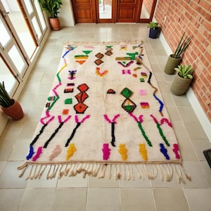 May include: A rectangular, off-white rug with a colorful geometric pattern. The design includes shapes in pink, green, orange, yellow, and blue. The rug has a fringe along the bottom edge and is placed on a tiled floor.