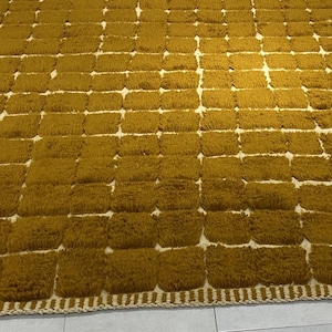 Large handmade Moroccan high-pile shag rug in saturated Thai curry mustard-gold, displayed vertically against a white brick wall. Surface features a tone-on-tone square grid pattern formed through pile variation — raised mustard squares separated by recessed ivory cream channel lines across the entire field. Dense, plush, curly wool pile texture throughout. Knotted braided ivory fringe along the top hem edge and long hand-tied ivory fringe along the bottom edge.