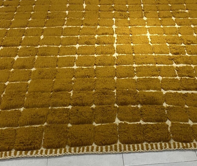Large handmade Moroccan high-pile shag rug in saturated Thai curry mustard-gold, displayed vertically against a white brick wall. Surface features a tone-on-tone square grid pattern formed through pile variation — raised mustard squares separated by recessed ivory cream channel lines across the entire field. Dense, plush, curly wool pile texture throughout. Knotted braided ivory fringe along the top hem edge and long hand-tied ivory fringe along the bottom edge.