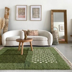 A plush handwoven Moroccan Berber rug in a rich moss green. The thick wool carpet features a solid dark green band near the top and a lighter mossy green base beautifully adorned with small, cream-colored geometric diamond motifs at the bottom. It is draped elegantly against a white painted brick wall next to a gold accent table with trailing ivy, perfectly capturing a warm, earthy, and organic modern interior design aesthetic.