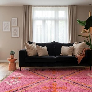 A plush, handwoven Moroccan Boujad rug featuring a vibrant hot pink base adorned with neon orange crosses, white diamonds, and green zigzag geometric patterns. The brightly colored wool carpet is draped elegantly against a white painted brick wall next to a gold accent table with pink pillar candles, perfectly capturing a playful maximalist and dopamine decor aesthetic.