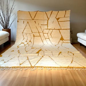 Handmade Moroccan rug in a soft ivory base with striking mustard yellow (Thai Curry) abstract lines. Woven by Berber artisans in pure wool, this rug blends tradition and modern style. Its geometric tribal pattern and warm tones make it perfect for living rooms, bedrooms, or creative interiors. Durable, eco-friendly, and soft underfoot, this ivory and mustard Berber carpet adds Moroccan charm and luxury to boho, modern, or eclectic home decor.