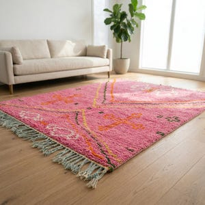 A plush, handwoven Moroccan Boujad rug featuring a vibrant hot pink base adorned with neon orange crosses, white diamonds, and green zigzag geometric patterns. The brightly colored wool carpet is draped elegantly against a white painted brick wall next to a gold accent table with pink pillar candles, perfectly capturing a playful maximalist and dopamine decor aesthetic.
