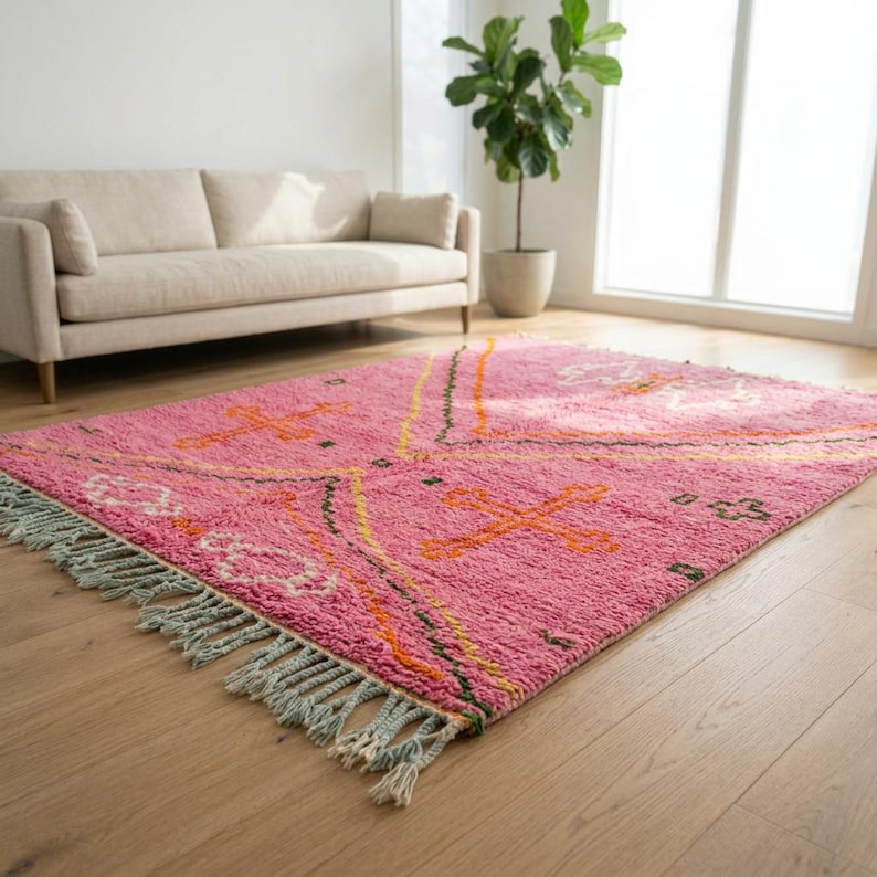 A plush, handwoven Moroccan Boujad rug featuring a vibrant hot pink base adorned with neon orange crosses, white diamonds, and green zigzag geometric patterns. The brightly colored wool carpet is draped elegantly against a white painted brick wall next to a gold accent table with pink pillar candles, perfectly capturing a playful maximalist and dopamine decor aesthetic.