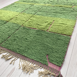 Puede incluir: Una alfombra rectangular con un diseño de patchwork en tonos verdes. La alfombra tiene un aspecto texturizado y peludo y está rematada con un borde rosa y verde y flecos beige.