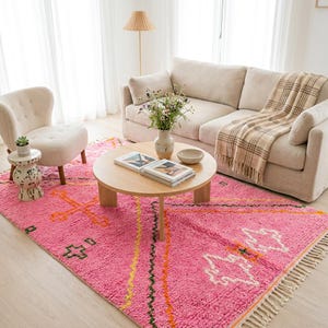 A plush, handwoven Moroccan Boujad rug featuring a vibrant hot pink base adorned with neon orange crosses, white diamonds, and green zigzag geometric patterns. The brightly colored wool carpet is draped elegantly against a white painted brick wall next to a gold accent table with pink pillar candles, perfectly capturing a playful maximalist and dopamine decor aesthetic.