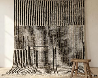May include: A large, black and white striped rug with a geometric design is hanging on a wall. The rug has a fringe at the bottom and is made of a thick, textured material. A small wooden stool sits in front of the rug.