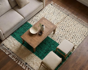 May include: A rectangular area rug featuring a cream-coloured section with small black flecks and a solid emerald green stripe, finished with knotted fringe on the ends. The rug is positioned under a wooden coffee table and two small stools.