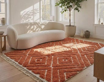May include: A rectangular area rug featuring a burnt orange and cream geometric diamond pattern with a high-pile texture and knotted fringe along the short edges. The rug is positioned on a light wood floor in front of a curved cream sofa.
