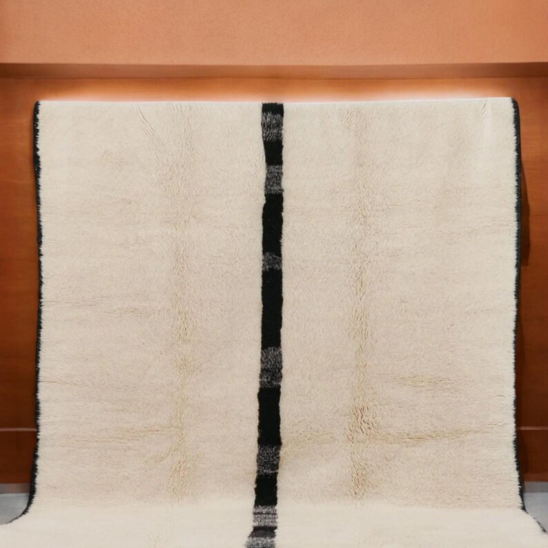 Handmade Moroccan wool rug featuring a minimalist black and white design. Ethically crafted by Berber artisans in Morocco using 100% natural wool. Thick, plush, and ultra-soft, this Beni Ourain-style carpet is perfect for modern, Scandinavian, and bohemian interiors. Ideal for living rooms, bedrooms, and luxury home decor. Available in custom sizes. Authentic tribal craftsmanship with eco-friendly materials. Free worldwide shipping with FedEx Express.
