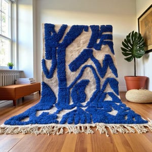 May include: A large, rectangular rug with a cream-colored background and vibrant blue abstract shapes. The rug has a thick, textured pile and a fringed edge. The rug is displayed on a hardwood floor.
