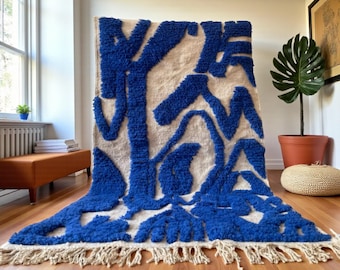 Puede incluir: Una alfombra rectangular grande con un fondo crema y formas abstractas azules vibrantes. La alfombra tiene un pelo grueso y texturizado y un borde con flecos. Se exhibe sobre un suelo de madera.