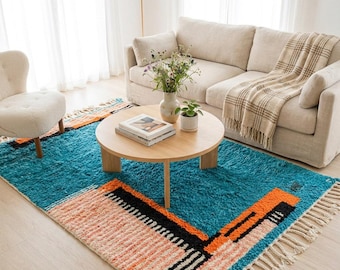 May include: A rectangular area rug featuring a vibrant teal base with geometric blocks of peach, orange, and black. The rug has a thick, shaggy texture and is finished with long, cream-coloured fringe tassels along the two shorter ends.