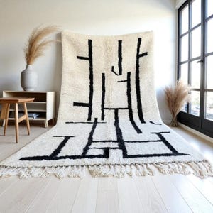 May include: An off-white rug with a striking black geometric pattern. The rug has a thick, textured pile and a fringe along the bottom. The abstract design features vertical and horizontal lines, creating a modern look. The rug is suitable for a living room.