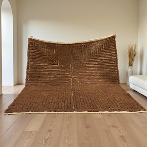 Pode incluir: Um tapete grande, castanho, texturizado com um design geométrico. O tapete tem uma pilha espessa e uma borda creme. O design apresenta uma série de linhas horizontais e verticais, criando um padrão semelhante a uma grade. O tapete é exibido em uma sala bem iluminada com piso de madeira.