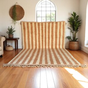 Handmade Moroccan striped wool rug in soft Cameo Rose and white tones, featuring traditional vertical patterns. Ideal for boho, Scandinavian, or modern interiors. Ethically woven by Berber artisans, this plush rug adds warmth, texture, and natural charm to your living room, bedroom, or reading nook. Custom sizes available. Perfect for neutral and pastel decor lovers.