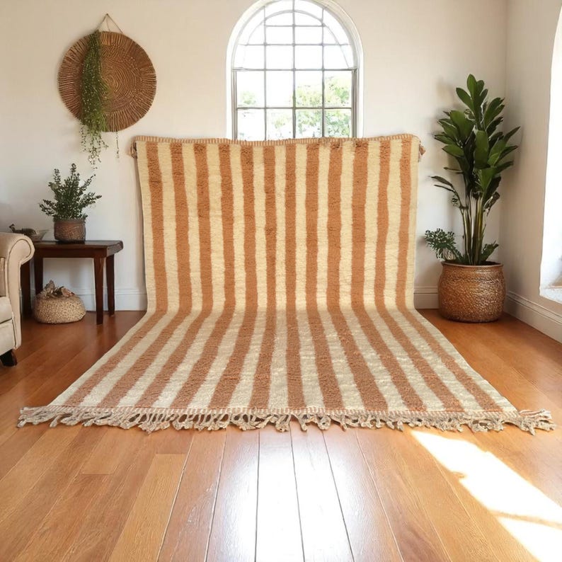 Handmade Moroccan striped wool rug in soft Cameo Rose and white tones, featuring traditional vertical patterns. Ideal for boho, Scandinavian, or modern interiors. Ethically woven by Berber artisans, this plush rug adds warmth, texture, and natural charm to your living room, bedroom, or reading nook. Custom sizes available. Perfect for neutral and pastel decor lovers.