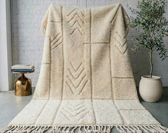May include: A cream-coloured, high-pile area rug featuring a geometric pattern of raised, textured chevron lines and rectangular sections. The rug is finished with a fringe of knotted tassels along the bottom edge, creating a soft, tactile surface.