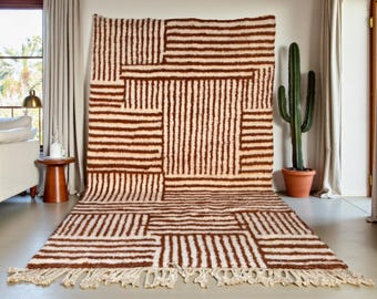 Puede incluir: Una alfombra rectangular grande con un patrón geométrico de rayas marrones sobre un fondo crema. La alfombra tiene un borde con flecos y se exhibe en una habitación con un cactus y cortinas. Adecuada para una sala de estar o dormitorio.