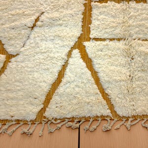 Handmade Moroccan rug in a soft ivory base with striking mustard yellow (Thai Curry) abstract lines. Woven by Berber artisans in pure wool, this rug blends tradition and modern style. Its geometric tribal pattern and warm tones make it perfect for living rooms, bedrooms, or creative interiors. Durable, eco-friendly, and soft underfoot, this ivory and mustard Berber carpet adds Moroccan charm and luxury to boho, modern, or eclectic home decor.