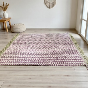 Handwoven Moroccan wool rug in wistful mauve and cream tones. Features a soft geometric pattern with textured pile and hand-knotted fringes. Perfect for living rooms, bedrooms, or boho interiors. Made from 100% natural wool by Berber artisans in Morocco. This handmade rug adds warmth, elegance, and Moroccan authenticity to modern or vintage-style spaces.