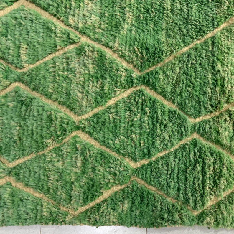 Green Moroccan wool rug with cream diamond lattice pattern, handmade in Beni Ourain style. Styled in a bright modern boho room with plants, rattan furniture, and natural light. The rug has a plush texture, tribal design, and fringe details, making it perfect for earthy, creative, or minimalist interiors. Crafted by Berber artisans, it's soft, thick, and ethically made using traditional weaving techniques.