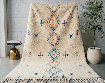 May include: A cream-coloured, high-pile area rug featuring a central vertical column of interconnected diamond shapes in orange, pink and teal. The rug is accented with small, scattered geometric motifs in matching colours and has a fringed edge.