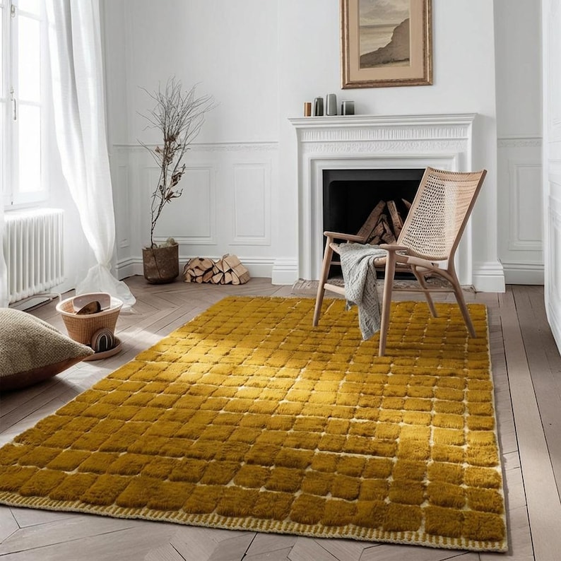 Large handmade Moroccan high-pile shag rug in saturated Thai curry mustard-gold, displayed vertically against a white brick wall. Surface features a tone-on-tone square grid pattern formed through pile variation — raised mustard squares separated by recessed ivory cream channel lines across the entire field. Dense, plush, curly wool pile texture throughout. Knotted braided ivory fringe along the top hem edge and long hand-tied ivory fringe along the bottom edge.