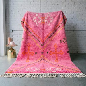 May include: A vibrant pink rug with a geometric pattern in orange, yellow, and white. The rug has a thick, textured pile and a fringe along the bottom edge. The design features cross-like shapes and diamond motifs.