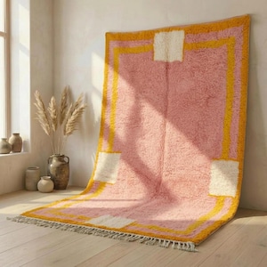 May include: A large, rectangular rug with a pink central field and a yellow border. The rug features white square accents and a fringed edge. The rug is displayed indoors, leaning against a white wall.
