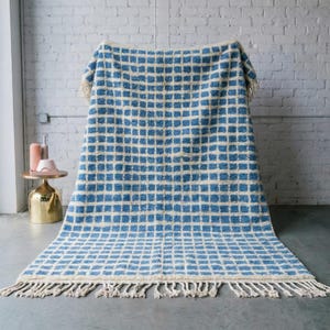 Peut inclure: Une couverture tissée bleue et crème avec un motif quadrillé. La couverture a une frange crème le long du bord inférieur. La couverture est drapée sur une surface contre un mur de briques blanches.