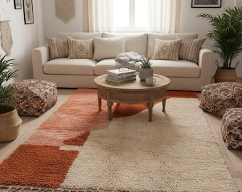 May include: A living room scene featuring a beige sofa with patterned pillows, a round wooden coffee table, and a large area rug. The rug has a geometric design in shades of orange and cream, with tassels along the edge. Decorative poufs and plants complete the cozy, inviting space.