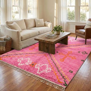 A plush, handwoven Moroccan Boujad rug featuring a vibrant hot pink base adorned with neon orange crosses, white diamonds, and green zigzag geometric patterns. The brightly colored wool carpet is draped elegantly against a white painted brick wall next to a gold accent table with pink pillar candles, perfectly capturing a playful maximalist and dopamine decor aesthetic.