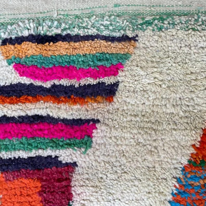 A vibrant, high-pile handmade Moroccan wool rug displayed in a bright industrial studio against a white brick wall. The rug features an eclectic, abstract tribal design with bold splashes of neon pink, teal green, orange, royal blue, and charcoal grey on a natural cream base. The patterns include geometric shapes, abstract figures, and spontaneous color blocking. The rug showcases a thick, plush shaggy texture and authentic hand-knotted cream tassels at the bottom.