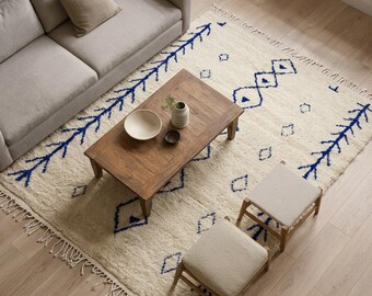 May include: A rectangular area rug featuring a cream-coloured base with blue geometric patterns and fringe edges. The rug is positioned under a wooden coffee table, two light-coloured upholstered stools, and a light grey sofa with a beige cushion.