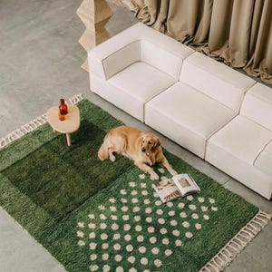 A plush handwoven Moroccan Berber rug in a rich moss green. The thick wool carpet features a solid dark green band near the top and a lighter mossy green base beautifully adorned with small, cream-colored geometric diamond motifs at the bottom. It is draped elegantly against a white painted brick wall next to a gold accent table with trailing ivy, perfectly capturing a warm, earthy, and organic modern interior design aesthetic.