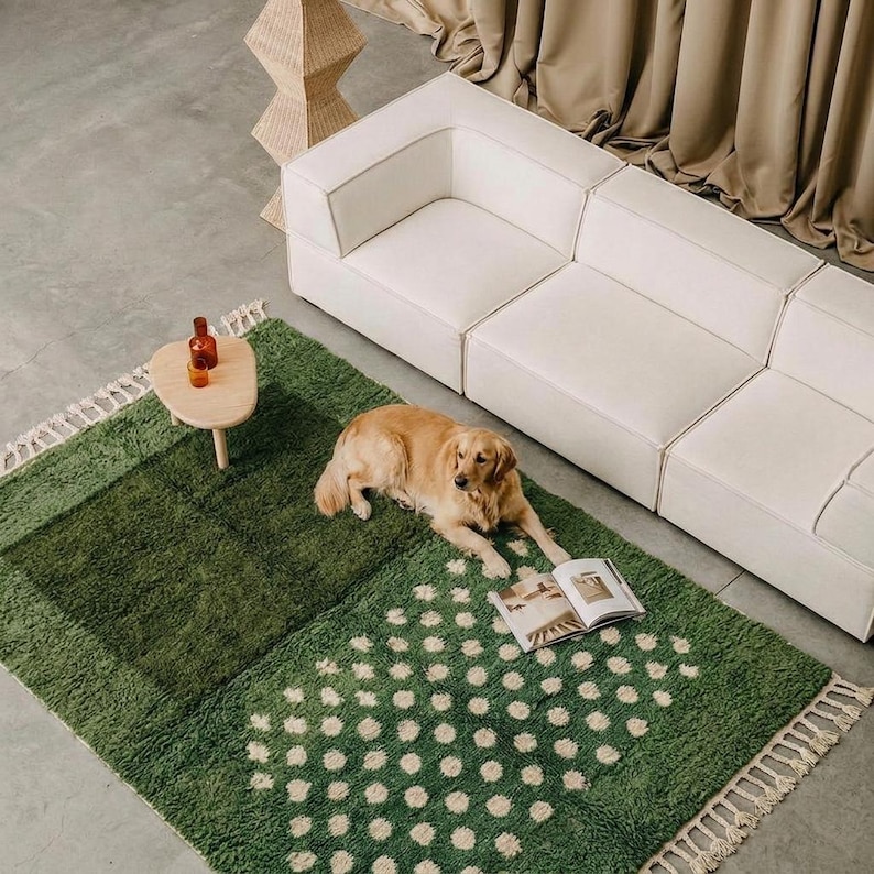 A plush handwoven Moroccan Berber rug in a rich moss green. The thick wool carpet features a solid dark green band near the top and a lighter mossy green base beautifully adorned with small, cream-colored geometric diamond motifs at the bottom. It is draped elegantly against a white painted brick wall next to a gold accent table with trailing ivy, perfectly capturing a warm, earthy, and organic modern interior design aesthetic.
