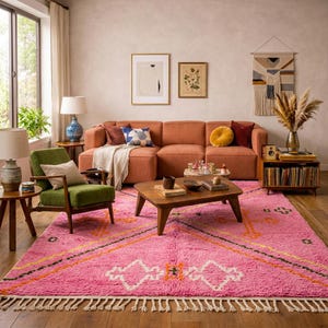 A plush, handwoven Moroccan Boujad rug featuring a vibrant hot pink base adorned with neon orange crosses, white diamonds, and green zigzag geometric patterns. The brightly colored wool carpet is draped elegantly against a white painted brick wall next to a gold accent table with pink pillar candles, perfectly capturing a playful maximalist and dopamine decor aesthetic.