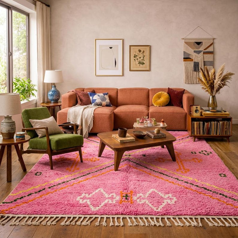 A plush, handwoven Moroccan Boujad rug featuring a vibrant hot pink base adorned with neon orange crosses, white diamonds, and green zigzag geometric patterns. The brightly colored wool carpet is draped elegantly against a white painted brick wall next to a gold accent table with pink pillar candles, perfectly capturing a playful maximalist and dopamine decor aesthetic.