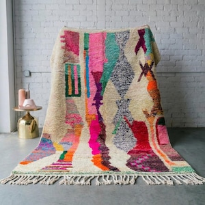 Op de afbeelding: Een rechthoekig tapijt met een crèmekleurige achtergrond en een levendig geometrisch ontwerp. Het tapijt heeft een verscheidenheid aan kleuren, waaronder roze, oranje, groen en blauw, met een gefranjerde rand. Het tapijt wordt getoond op een grijze vloer tegen een witte bakstenen muur.