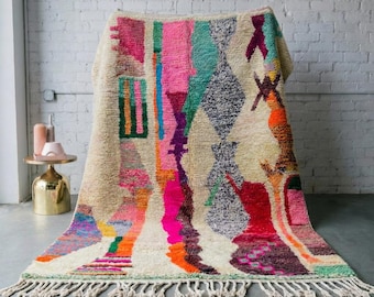 Può includere: Un tappeto rettangolare dai colori vivaci con uno sfondo color crema e un design geometrico. Il tappeto presenta una varietà di colori, tra cui rosa, arancione, verde e blu, con un bordo a frange. Il tappeto è esposto su un pavimento grigio contro un muro di mattoni bianchi.