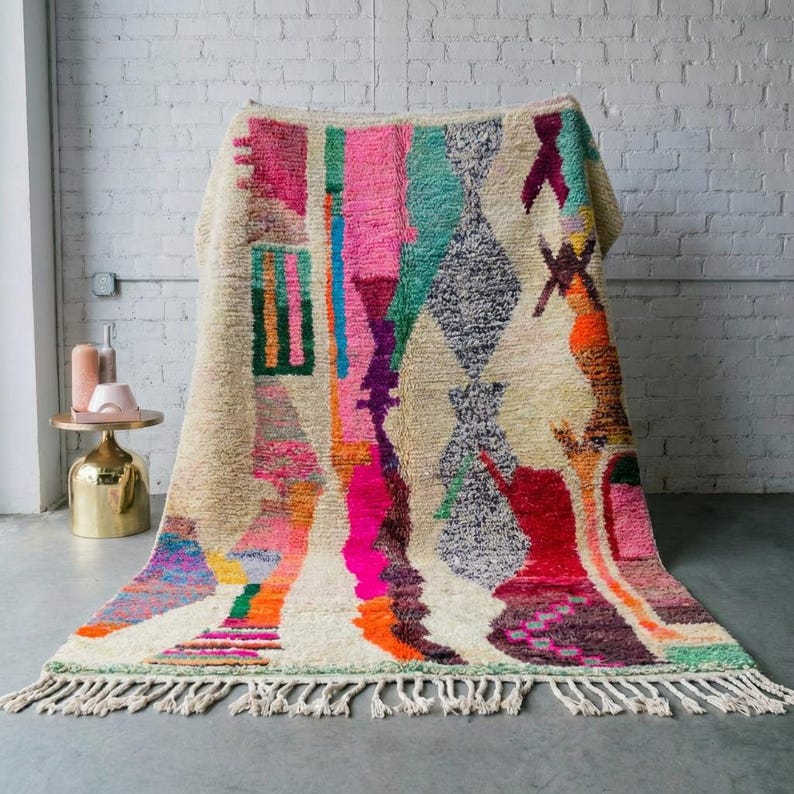 A vibrant, high-pile handmade Moroccan wool rug displayed in a bright industrial studio against a white brick wall. The rug features an eclectic, abstract tribal design with bold splashes of neon pink, teal green, orange, royal blue, and charcoal grey on a natural cream base. The patterns include geometric shapes, abstract figures, and spontaneous color blocking. The rug showcases a thick, plush shaggy texture and authentic hand-knotted cream tassels at the bottom.