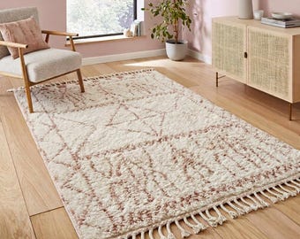 Può includere: Un tappeto rettangolare con base color crema e un motivo geometrico in rosa tenue. Il tappeto presenta una consistenza a pelo folto e morbido, rifinito con lunghe frange annodate sui bordi più corti, che conferiscono un aspetto strutturato.